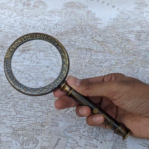 May include: A vintage brass magnifying glass with the text "HENRY HUGHES & SON LTD. LONDON 1941" around the rim. The magnifying glass is held over a detailed map, showcasing its use for examining maps and documents.