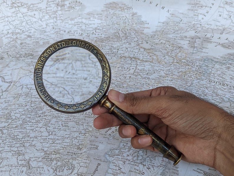 May include: A vintage brass magnifying glass with the text "HENRY HUGHES & SON LTD. LONDON 1941" around the rim. The magnifying glass is held over a detailed map, showcasing its use for examining maps and documents.