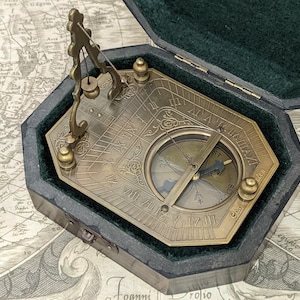 May include: A vintage brass sundial compass in a dark green velvet-lined case. The compass features Roman numerals and a needle. The case is octagonal and rests on a map.