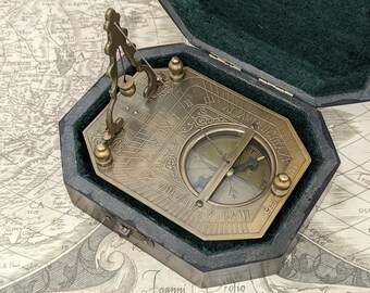 May include: A vintage brass sundial compass in a dark green velvet-lined case. The compass features Roman numerals and a needle. The case is octagonal and rests on a map.