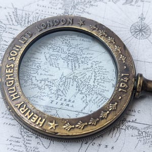 May include: Antique brass magnifying glass with a dark handle, resting on a vintage map. The magnifying glass has the text "HENRY HUGHES SON LTD. LONDON 1941" around the rim. The map is in shades of gray and white.