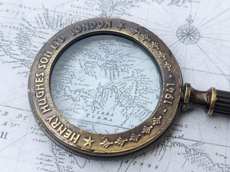 May include: Antique brass magnifying glass with a dark handle, resting on a vintage map. The magnifying glass has the text "HENRY HUGHES SON LTD. LONDON 1941" around the rim. The map is in shades of gray and white.