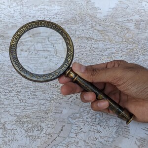 May include: A vintage brass magnifying glass with the text "HENRY HUGHESSON LTD. LONDON 1941" around the rim. The magnifying glass is held over a detailed map, showcasing its use for close inspection of cartography and historical documents.