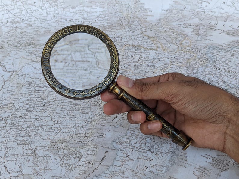 May include: A vintage brass magnifying glass with the text "HENRY HUGHESSON LTD. LONDON 1941" around the rim. The magnifying glass is held over a detailed map, showcasing its use for close inspection of cartography and historical documents.