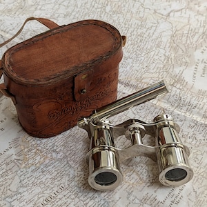 May include: Vintage silver opera glasses with a brown leather case. The case has a handle and is embossed with text. The glasses are resting on a vintage map.