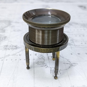 May include: A vintage brass magnifying glass with a round lens and a detailed, ridged body. The base is supported by three slender legs and features the engraved text "ROSS LONDON". The magnifying glass is a classic antique.