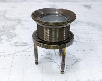 May include: A vintage brass magnifying glass with a round lens and a detailed, ridged body. The base is supported by three slender legs and features the engraved text "ROSS LONDON". The magnifying glass is a classic antique.