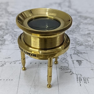 May include: A brass tripod magnifier with a circular lens and a knurled edge. The base features three slender legs and the text ROSS LONDON engraved on the side. The metallic finish is polished and reflective, showcasing a cylindrical body design.
