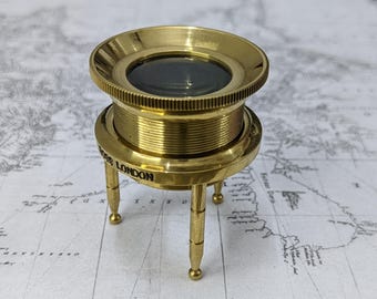 May include: A brass tripod magnifier with a circular lens and a knurled edge. The base features three slender legs and the text ROSS LONDON engraved on the side. The metallic finish is polished and reflective, showcasing a cylindrical body design.