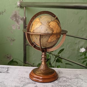 Pode incluir: Um globo de estilo vintage com uma esfera creme e um mapa detalhado. O globo é mantido dentro de uma estrutura de metal cor de bronze e montado em uma base de madeira. A base tem um design ornamentado. É um item decorativo.
