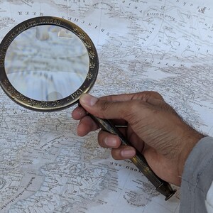 May include: A vintage brass magnifying glass with a detailed floral design around the rim and handle. The text "HENRY HUGHES & SON LTD LONDON - 1941" is visible on the rim. The magnifying glass is held over a detailed antique map.