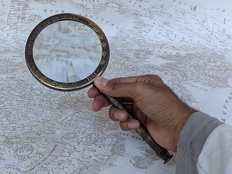 May include: A vintage brass magnifying glass with a detailed floral design around the rim and handle. The text "HENRY HUGHES & SON LTD LONDON - 1941" is visible on the rim. The magnifying glass is held over a detailed antique map.
