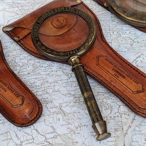 May include: Antique brass magnifying glass with a leather case. The magnifying glass has a detailed handle and a round lens. The leather case is brown and embossed with the text "HENRY HUGHES SONS LTD LONDON 1941".