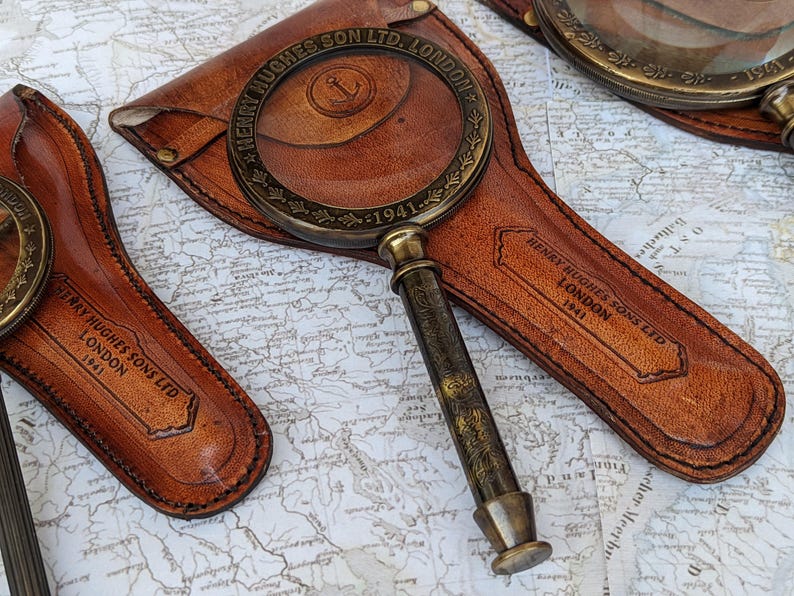 May include: Antique brass magnifying glass with a leather case. The magnifying glass has a detailed handle and a round lens. The leather case is brown and embossed with the text "HENRY HUGHES SONS LTD LONDON 1941".