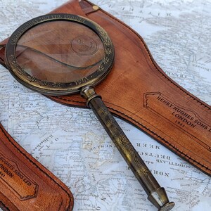 May include: Antique brass magnifying glass with a leather case. The magnifying glass has a detailed handle and a decorative rim. The leather case is embossed with the text "HENRY HUGHES SONS LTD LONDON 1941".