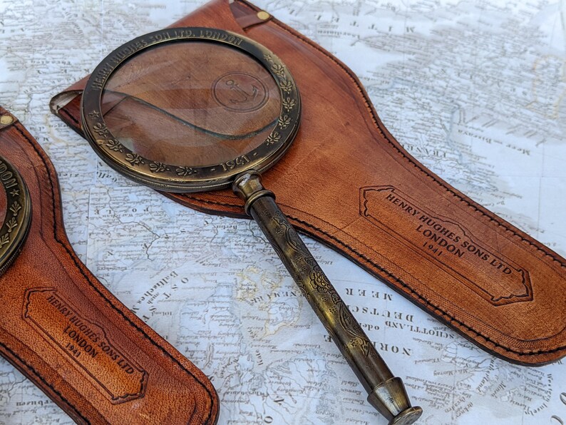 May include: Antique brass magnifying glass with a leather case. The magnifying glass has a detailed handle and a decorative rim. The leather case is embossed with the text "HENRY HUGHES SONS LTD LONDON 1941".