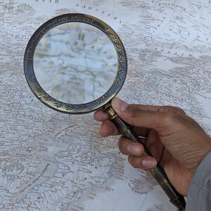 May include: A vintage brass magnifying glass with a detailed handle and a circular frame. The frame is engraved with text and floral patterns. The magnifying glass is held over a detailed map.