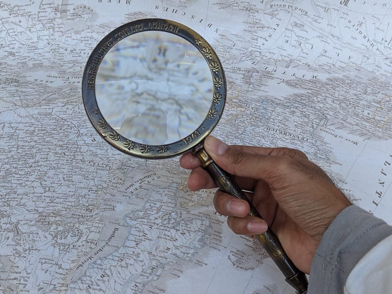 May include: A vintage brass magnifying glass with a detailed handle and a circular frame. The frame is engraved with text and floral patterns. The magnifying glass is held over a detailed map.