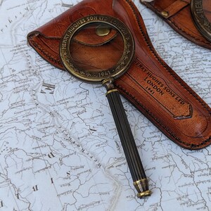 May include: A vintage brass and black magnifying glass with a leather case. The magnifying glass has a detailed handle and a circular lens. The leather case is brown and embossed with the text "HENRY HUGHES SONS LTD LONDON 1941". The items are on a map.