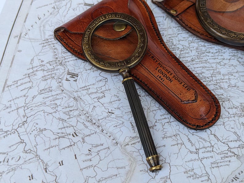 May include: A vintage brass and black magnifying glass with a leather case. The magnifying glass has a detailed handle and a circular lens. The leather case is brown and embossed with the text "HENRY HUGHES SONS LTD LONDON 1941". The items are on a map.