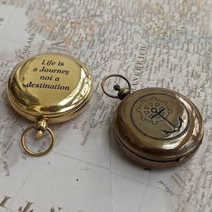 May include: Two round brass compasses with circular loops. One compass features the engraved text "Life is a Journey not a destination" on its polished surface, while the other displays an etched nautical anchor and ship wheel design.