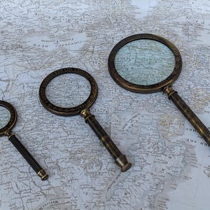 May include: Three antique brass magnifying glasses of varying sizes rest on a vintage map. The magnifying glasses have ornate detailing around the lenses and textured handles. The map provides a historical backdrop, enhancing the vintage aesthetic.