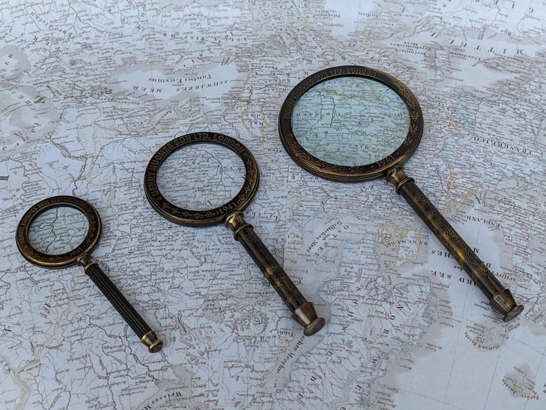 May include: Three antique brass magnifying glasses of varying sizes rest on a vintage map. The magnifying glasses have ornate detailing around the lenses and textured handles. The map provides a historical backdrop, enhancing the vintage aesthetic.
