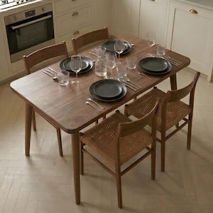 Può includere: Una sala da pranzo con un tavolo in legno e quattro sedie. Il tavolo è apparecchiato con piatti neri, posate e bicchieri. Le sedie hanno sedute intrecciate. La cucina ha mobili bianchi e una finestra.