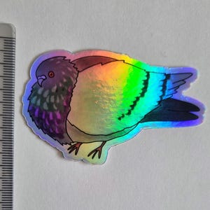 May include: Holographic sticker of a pigeon with iridescent wings. The bird's body features shades of purple, brown, and grey. The sticker is approximately 5 cm long, with a ruler in the background for scale. The sticker has a glossy finish.
