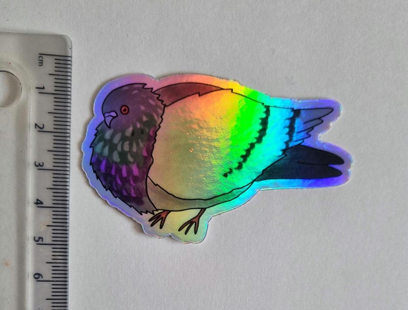 May include: Holographic sticker of a pigeon with iridescent wings. The bird's body features shades of purple, brown, and grey. The sticker is approximately 5 cm long, with a ruler in the background for scale. The sticker has a glossy finish.