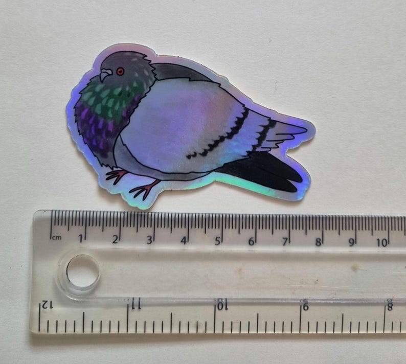May include: A holographic sticker of a pigeon. The sticker features a detailed illustration of the bird with iridescent colors, including purple, green, and gray. The sticker is approximately 7.6 cm long, as measured by the ruler in the image.