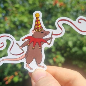 May include: A sticker of a cartoon rat in a jester's outfit, with a red and gold hat and collar. It holds two small sticks and a red ribbon. The background is a blurred green.