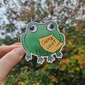 May include: A green frog sticker with large eyes and a yellow sticky note that reads "DUMB IDIOT." The sticker is held in a hand against a blurred background of autumn leaves. The frog is a cartoon illustration.