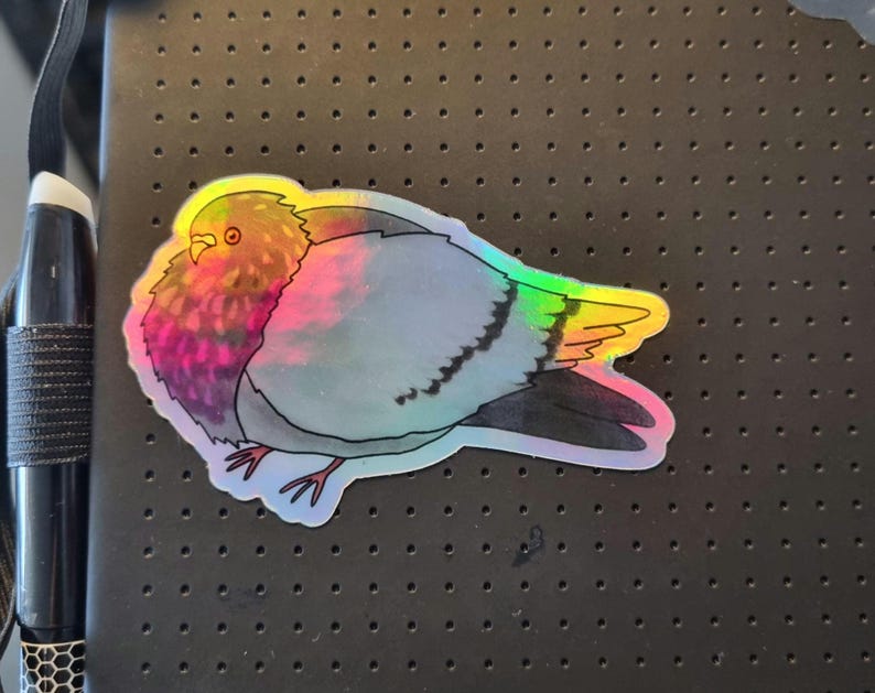 May include: A holographic sticker of a pigeon. The bird has a grey body, iridescent wings, and a colourful chest with pink, purple, and orange hues. The sticker has a white border and is on a black surface.
