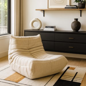 Op de afbeelding: Een crèmekleurige, gewatteerde loungestoel met een laag profiel. De stoel staat op een patroonkleed met zwarte, bruine en witte geometrische vormen. De kamer heeft een zwarte kast, een plank met decoratie en een raam.