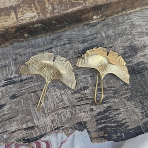 Puede incluir: Dos ganchos de pared de metal dorado con forma de hoja de Ginkgo. Los ganchos están detallados con un patrón en forma de abanico y tienen un tallo curvado para colgar. Se muestran sobre una superficie de madera envejecida.