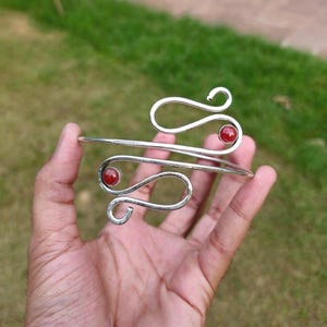 Può includere: Un bracciale a fascia in metallo color argento caratterizzato da un design a filo aperto e sinuoso. Il bracciale è impreziosito da due piccole pietre rosse rotonde e levigate, fissate alle estremità dei fili curvi per un aspetto simmetrico.