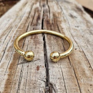 May include: A polished gold-tone metal cuff bracelet featuring a smooth, open-ended circular band design. The two ends of the bracelet terminate in rounded, spherical ball accents that face each other, creating a simple and continuous metallic loop.