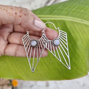 May include: Silver-toned geometric earrings with a triangular design. Each earring features a central, round, light-colored stone and a series of radiating lines. The earrings are held against a green leaf background.