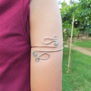May include: A silver-toned metal arm cuff featuring a wire-wrapped design with two swirling ends. Each end is accented with a small, round, translucent white gemstone. The cuff is open-ended, allowing it to wrap around the upper arm securely.
