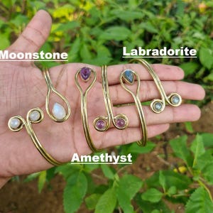 Peut inclure: Quatre bracelets manchettes en métal doré ornés de diverses pierres précieuses. Un bracelet possède une pierre de lune, un une améthyste, un une labradorite et un une petite pierre ronde. Le texte Moonstone, Labradorite et Amethyst est visible.