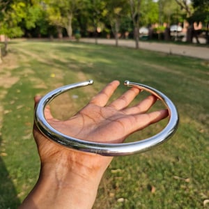 May include: A polished silver-tone metal torque necklace featuring a smooth, circular band design with two small spherical end caps. The rigid, open-ended collar is crafted from a reflective, solid material that maintains a consistent, rounded shape.