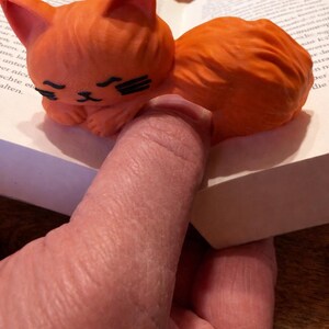 Könnte beinhalten: Orangefarbene Katzenfigur mit schwarzen Akzenten, die auf einem Buch liegt. Die Katze ist zusammengerollt, die Augen geschlossen. Die Figur ist einfarbig und scheint aus einem harten Material zu bestehen.