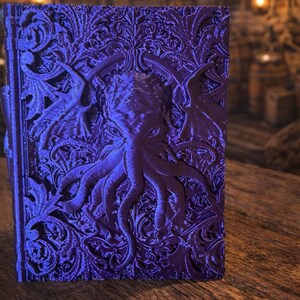 Könnte beinhalten: Ein detailliertes, lila Buch mit einem geprägten Design, das eine oktopusartige Kreatur und verzierte Blumenmuster zeigt. Das Buchcover trägt den Text "FATESEND" und "YEAR OF THE SNAKE" mit dem Jahr "2025" in einem kreisförmigen Emblem.