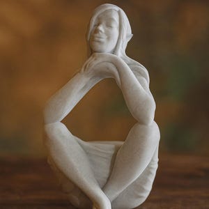 Può includere: Una serena scultura bianca di una figura seduta con le gambe incrociate e le braccia appoggiate sulle ginocchia. La figura ha gli occhi chiusi e un'espressione gentile. La scultura è su una superficie di legno.