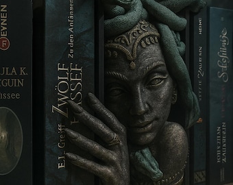 May include: A dark-toned bookend featuring a sculpted face with snakes for hair, positioned between two black books. The figure has a hand resting against the side of the book, wearing a ring. The book spine displays the text Zwölf Wasser.