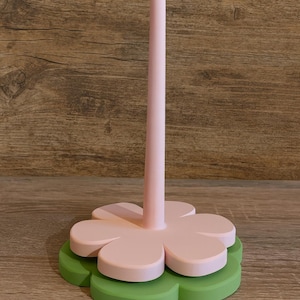 Könnte beinhalten: Ein Papiertuchhalter aus Holz in Blumenform. Die Basis ist grün, darüber eine rosa Blütenform. Ein hoher, rosa zylindrischer Pfosten erhebt sich aus der Mitte. Der Hintergrund ist eine Holzoberfläche.