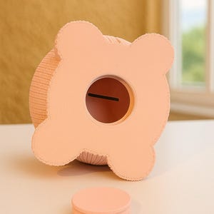 May include: A peach-colored piggy bank with a round coin slot and a stack of matching coasters. The bank has a textured, knitted body and a four-lobed top. The design is simple and modern, suitable for a nursery or a child's room.