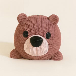 May include: A brown teddy bear-shaped decorative object with a knitted texture. The bear has black eyes and a pink nose. The object is sitting on a white surface. The bear is a cute and whimsical item.