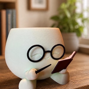 May include: A pale green, rounded planter shaped like a character with a smooth finish. It features black circular glasses, a small smiling mouth, and two small feet. The character holds a small black pen and an open dark brown book.